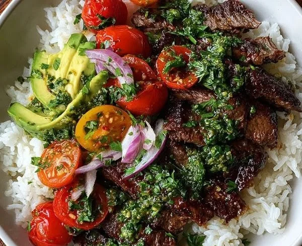 Skirt steak rice bowl topped with chimichurri sauce and fresh vegetables